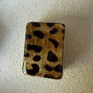 May include: A rectangular tile featuring a glossy finish with a gold and black leopard print pattern. The surface has a smooth, reflective texture with distinct dark spots set against a metallic gold background, creating a clear, repeating animal-inspired design.