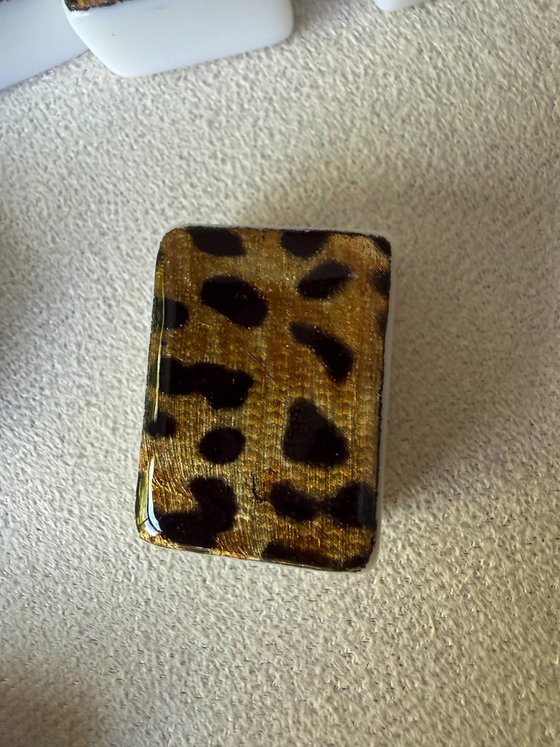 May include: A rectangular tile featuring a glossy finish with a gold and black leopard print pattern. The surface has a smooth, reflective texture with distinct dark spots set against a metallic gold background, creating a clear, repeating animal-inspired design.