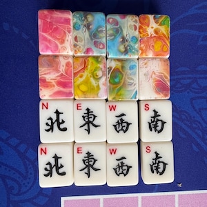 May include: A collection of colorful Mahjong tiles. The top row features tiles with abstract patterns in pink, blue, and orange. The middle row has green and pink tiles. The bottom two rows display white tiles with black Chinese characters and the letters N, E, W, and S.