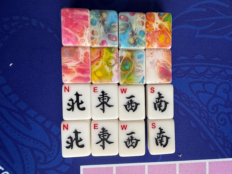 May include: A collection of colorful Mahjong tiles. The top row features tiles with abstract patterns in pink, blue, and orange. The middle row has green and pink tiles. The bottom two rows display white tiles with black Chinese characters and the letters N, E, W, and S.
