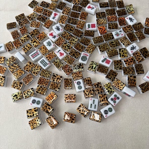 May include: A collection of rectangular Mahjong tiles scattered across a surface. The tiles feature a white base with a black and brown leopard print pattern on the back, while the front faces display various traditional symbols, numbers, and characters.