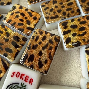 May include: A collection of rectangular mahjong tiles featuring a gold and black leopard print pattern on the top surface. One tile displays the word JOKER in red text above a green circular graphic, while another shows a red number seven.