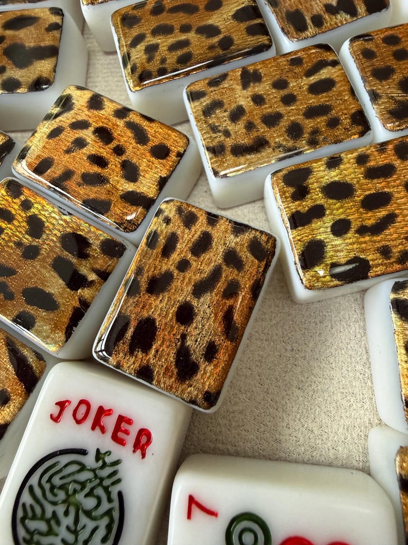 May include: A collection of rectangular mahjong tiles featuring a gold and black leopard print pattern on the top surface. One tile displays the word JOKER in red text above a green circular graphic, while another shows a red number seven.