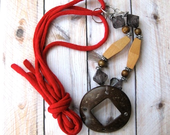 May include: A handmade necklace featuring a large, dark brown, circular pendant with a diamond-shaped cutout. The necklace includes wooden and gray bead accents, and a bright red fabric cord. The pendant is approximately 3 inches in diameter.
