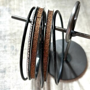 May include: A set of five thin bangles displayed on a metal stand. The collection includes two plain black bangles, one black bangle with a textured silver-tone edge, and two copper-colored bangles with a fine, sparkling glitter finish throughout.