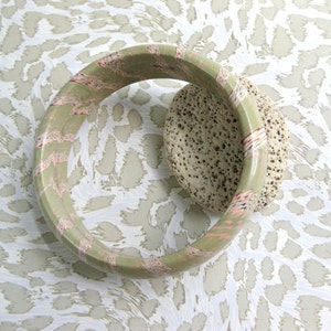 May include: A green and pink bangle bracelet with a textured surface. The bracelet is on a white and brown patterned background.