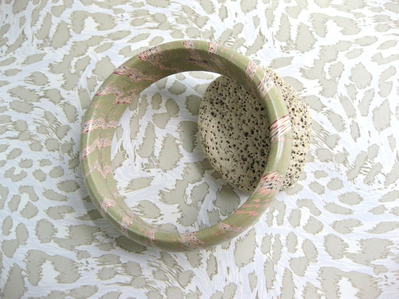 May include: A green and pink bangle bracelet with a textured surface. The bracelet is on a white and brown patterned background.