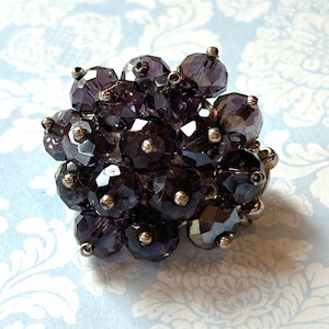 May include: A cluster ring featuring faceted, dark purple beads with silver-tone metal accents. The beads are arranged in a floral or spherical shape. The ring is set against a light blue background with a floral pattern.