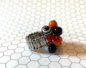 May include: A beaded ring with a silver stretch band. The ring features a cluster of beads in orange, black, and white, with a few red crystal beads. The ring is set against a white and gray hexagonal pattern.