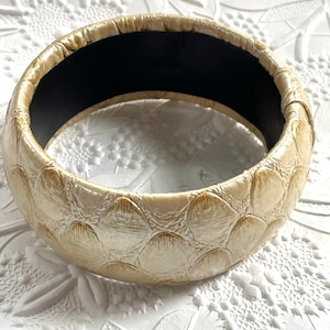 May include: A wide, cream-colored bangle bracelet with a textured, alligator-skin-like pattern. The inside of the bracelet is lined with black fabric.
