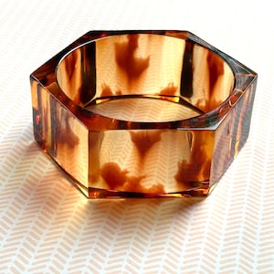 May include: A hexagonal bangle bracelet with a tortoiseshell pattern. The bracelet is made of a translucent material, showcasing shades of brown and amber. The geometric design adds a modern touch to this accessory.