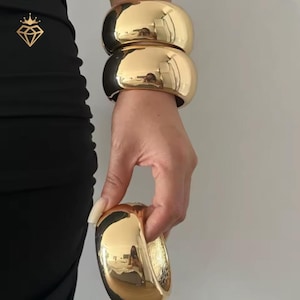 May include: Three thick, polished gold-tone metal bangle bracelets with a smooth, reflective surface. Two of the bangles are stacked together, while the third is held separately, showcasing a rounded, chunky design with a high-shine finish.