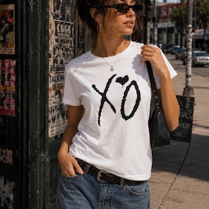 May include: A white short-sleeved t-shirt featuring a black graphic print of the letters XO with a small heart above the O. The shirt is worn with blue denim jeans, a black leather belt, a black shoulder bag, and dark-tinted sunglasses.