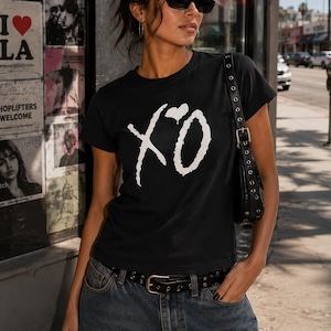 May include: A black short-sleeve t-shirt featuring a white graphic logo of the letters XO with a small heart above the O. The shirt is paired with blue denim jeans and a black leather belt with a silver buckle, along with a black shoulder bag.