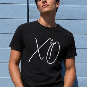 May include: A black short-sleeved t-shirt featuring the white text XO in a cursive script font. A small white heart outline is positioned above the letter O. The shirt has a crew neckline and is made of a solid black fabric with white graphic print.