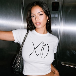 May include: A white short-sleeved crop top featuring the black text XO with a small heart symbol above the O. The garment is paired with blue denim jeans and a black shoulder bag with a silver chain strap, creating a casual look.