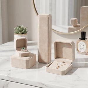 May include: Beige velvet jewelry boxes in various shapes and sizes. One box holds a ring with a green stone, another a necklace with a pendant. A succulent plant and perfume bottle are also visible.