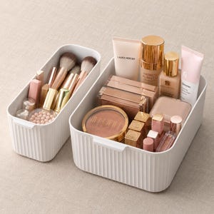 May include: Two white plastic storage bins with vertical ribbed sides. The smaller bin holds makeup brushes and small cosmetic items. The larger bin contains various beauty products including tubes, compacts, and lipsticks with visible brand labels.