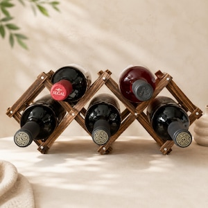 May include: A dark brown wooden accordion-style wine rack holding five glass wine bottles. The rack features a crisscross lattice design with metal rivets at each joint. One bottle cap is red with the text SEGAL, while others are black or grey.