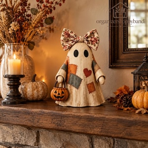 May include: A decorative ghost figurine featuring a cream-colored fabric body with stitched-on patches in muted tones, a red heart accent, and a large floral print bow on top. The ghost holds a small orange jack-o'-lantern bucket in one hand.