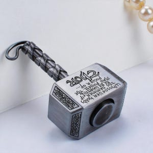 Puede incluir: Un martillo de Thor plateado con un mango trenzado y la palabra "THOR" en la parte superior. El martillo tiene un texto grabado que dice "Quien empuñe este martillo, si es digno, poseerá el poder de..."