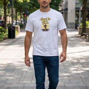 Puede incluir: Una camiseta blanca de manga corta con un estampado gr&aacute;fico que incluye el texto Saved by God's Grace, una cruz de madera marr&oacute;n, una forma de gota amarilla y el texto EPHESIANS 2:8, combinada con vaqueros azul oscuro y zapatillas grises.