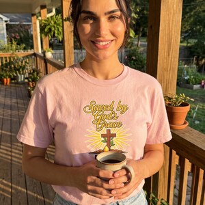 Puede incluir: Una camiseta de manga corta de color rosa claro con el texto "Saved by God's Grace" en una fuente cursiva amarilla sobre un gr&aacute;fico de una cruz marr&oacute;n, un peque&ntilde;o brote verde y un sol amarillo, combinada con unos vaqueros azul claro.