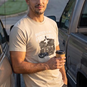 Puede incluir: Una camiseta beige de manga corta con un estampado gr&aacute;fico de una radio antigua y una cruz. El texto PRAYER IS MY FIRST CALL est&aacute; impreso en blanco sobre la ilustraci&oacute;n. La camiseta tiene cuello redondo y un corte est&aacute;ndar de algod&oacute;n.