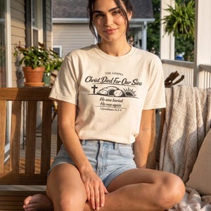 May include: A cream-coloured short-sleeve t-shirt featuring black text that reads THE GOSPEL Christ Died For Our Sins, He was buried, He rose again, 1 Corinthians 15:3-4, accompanied by a graphic of a cross, an open tomb, and a rising sun.