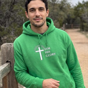 May include: A bright green hooded sweatshirt featuring a white graphic of a cross with radiating lines above it and the text "FOR HIS GLORY" printed in white. The pullover includes a front kangaroo pocket, long sleeves, and a drawstring hood.