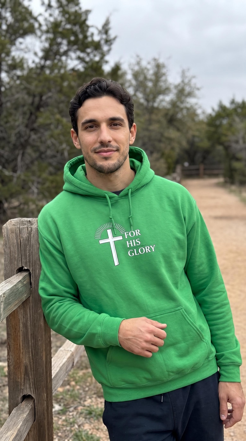 May include: A bright green hooded sweatshirt featuring a white graphic of a cross with radiating lines above it and the text "FOR HIS GLORY" printed in white. The pullover includes a front kangaroo pocket, long sleeves, and a drawstring hood.