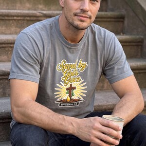 Puede incluir: Una camiseta gris de manga corta con un estampado gr&aacute;fico que incluye el texto "Saved by God's Grace" en letra cursiva amarilla, una cruz marr&oacute;n, una planta verde, una gota dorada y el texto "EPHESIANS 2:8" en un recuadro negro.