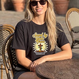 Puede incluir: Una camiseta negra de manga corta con un estampado gr&aacute;fico que incluye el texto "Saved by God's Grace" en letra amarilla, una cruz de madera marr&oacute;n, un brote verde, una gota amarilla y el texto "EPHESIANS 2:6" sobre un fondo blanco.