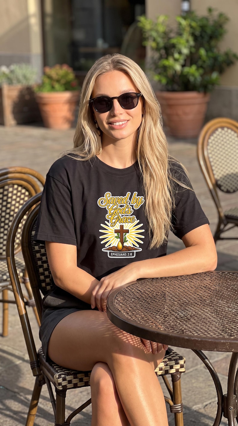 Puede incluir: Una camiseta negra de manga corta con un estampado gr&aacute;fico que incluye el texto "Saved by God's Grace" en letra amarilla, una cruz de madera marr&oacute;n, un brote verde, una gota amarilla y el texto "EPHESIANS 2:6" sobre un fondo blanco.