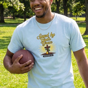 Puede incluir: Una camiseta de manga corta de color azul claro con un estampado gr&aacute;fico que incluye el texto "Saved by God's Grace" en letra amarilla, una cruz marr&oacute;n, un sol amarillo y una banda negra con el texto blanco "EPHESIANS 2:8".