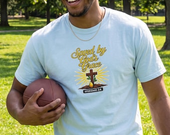 Könnte beinhalten: Ein hellblaues Kurzarm-T-Shirt mit einem Grafikdruck, der den Text "Saved by God's Grace" in gelber Schreibschrift, ein braunes Kreuz, einen gelben Sonnenstrahl und ein schwarzes Banner mit dem weißen Text "EPHESIANS 2:8" zeigt.