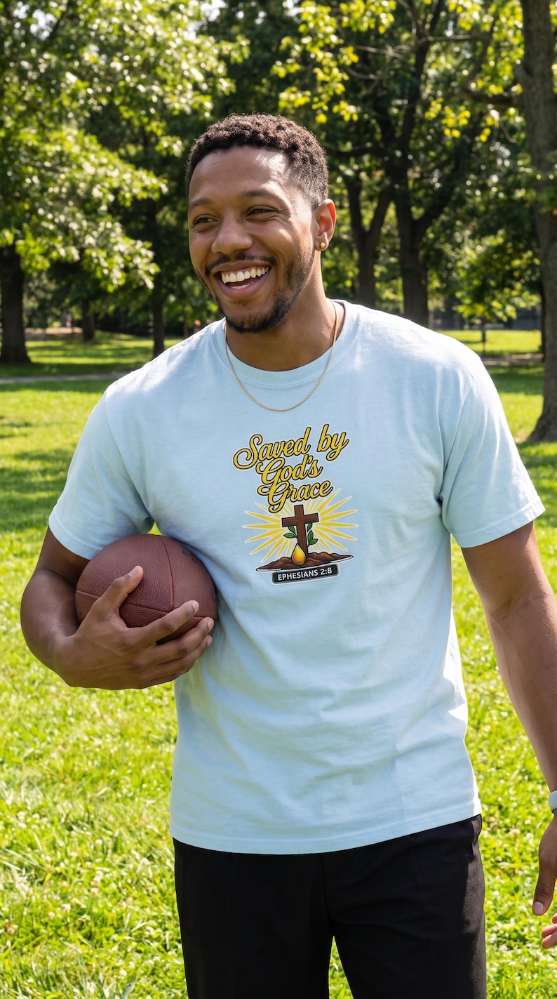 Puede incluir: Una camiseta de manga corta de color azul claro con un estampado gr&aacute;fico que incluye el texto "Saved by God's Grace" en letra amarilla, una cruz marr&oacute;n, un sol amarillo y una banda negra con el texto blanco "EPHESIANS 2:8".