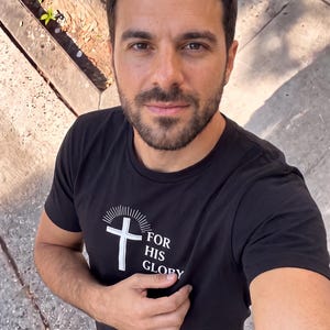 Puede incluir: Una camiseta negra de manga corta con un gráfico blanco de una cruz con líneas radiantes encima y el texto "FOR HIS GLORY" impreso en el pecho. La camiseta tiene un cuello redondo y está confeccionada en tela negra sólida con tinta blanca.