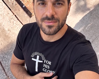 May include: A black short-sleeved t-shirt featuring a white graphic of a cross with radiating lines above it and the text "FOR HIS GLORY" printed on the chest. The shirt has a crew neckline and is made of a solid black fabric with white ink.