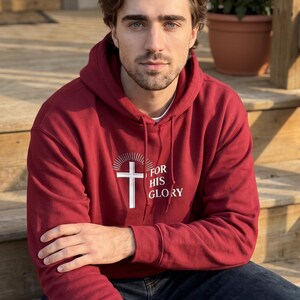 May include: A maroon hooded sweatshirt featuring a white graphic of a cross with radiating lines above it and the text FOR HIS GLORY printed to the right of the cross. The hoodie has long sleeves, a front pocket, and matching maroon drawstrings.