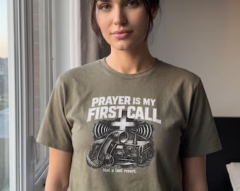 Könnte beinhalten: Ein olivgrünes Kurzarm-T-Shirt mit einer Grafik eines alten Funkgeräts, versehen mit dem Text PRAYER IS MY FIRST CALL und Not a last resort. Das Design zeigt ein Kreuzsymbol über dem Funkgerät sowie stilisierte Signalwellen.