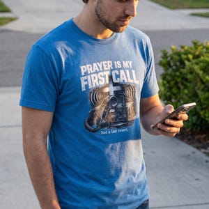 Puede incluir: Una camiseta azul de manga corta con un gr&aacute;fico de una radio antigua y una cruz. El texto dice PRAYER IS MY FIRST CALL y Not a last resort. El dise&ntilde;o est&aacute; impreso en tinta blanca y negra en la parte delantera de la camiseta de algod&oacute;n.