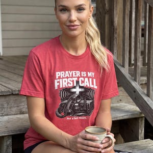 Puede incluir: Una camiseta roja de manga corta con un gr&aacute;fico de una radio y una cruz, junto al texto en blanco PRAYER IS MY FIRST CALL y Not a last resort. La prenda cuenta con cuello redondo, una textura jaspeada y una detallada ilustraci&oacute;n negra.