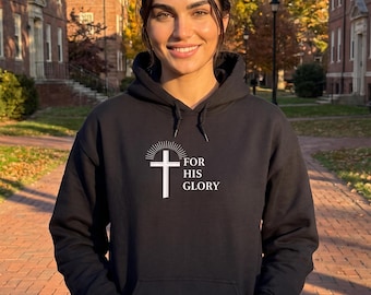 Könnte beinhalten: Ein schwarzer Kapuzenpullover mit einer weißen Grafik eines Kreuzes mit darüber liegenden Strahlen und dem Text FOR HIS GLORY rechts neben dem Kreuz. Das Kleidungsstück verfügt über eine Kängurutasche vorne und passende schwarze Kordelzüge.