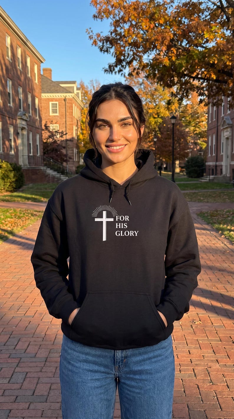 May include: A black hooded sweatshirt featuring a white graphic of a cross with radiating lines above it and the text FOR HIS GLORY printed to the right of the cross. The garment includes a front kangaroo pocket and matching black drawstrings.
