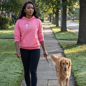May include: A bright pink hooded sweatshirt featuring a white cross graphic and the text "FOR HIS GLORY" on the chest, paired with black leggings and black athletic sneakers, while holding a tan leather leash attached to a golden retriever.