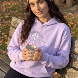 May include: A light purple hooded sweatshirt featuring a white cross graphic with sunburst rays and the text FOR HIS GLORY printed on the chest. The hoodie includes a front kangaroo pocket and long sleeves, paired with a grey floral patterned headscarf.
