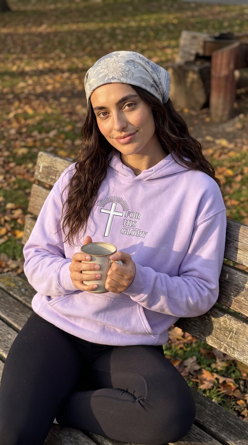 May include: A light purple hooded sweatshirt featuring a white cross graphic with sunburst rays and the text FOR HIS GLORY printed on the chest. The hoodie includes a front kangaroo pocket and long sleeves, paired with a grey floral patterned headscarf.