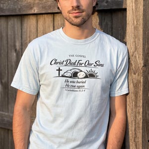 Puede incluir: Una camiseta de manga corta de color azul claro con texto negro que dice THE GOSPEL Christ Died For Our Sins, seguido de un gráfico de una cruz, una tumba abierta y un amanecer, con el texto He was buried He rose again 1 Corinthians 15:3-4.