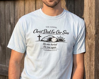 May include: A light blue short-sleeve t-shirt featuring black text that reads THE GOSPEL Christ Died For Our Sins, followed by a graphic of a cross, an open tomb, and a sunrise, with the text He was buried He rose again 1 Corinthians 15:3-4.